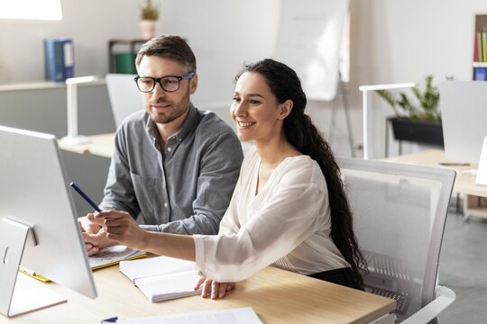 Smiling Hispanic Woman Assisting Caucasian Male Colleague Using Computer While Collaborating On Project At Office, Engaging In Professional Work Environment With Team Spirit.