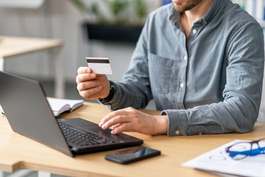 Focused Man Holding Credit Card While Working On Laptop At Office Desk. Engaged In Online Payment Process, Modern Work Environment. Business Concept.