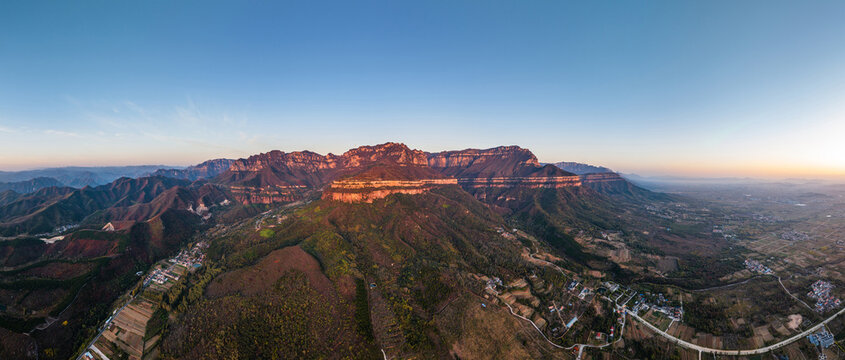 Aerial wide panorama shows sunlit layered mountain ranges, surrounding valleys, rural settlements and clear sky at dawn.