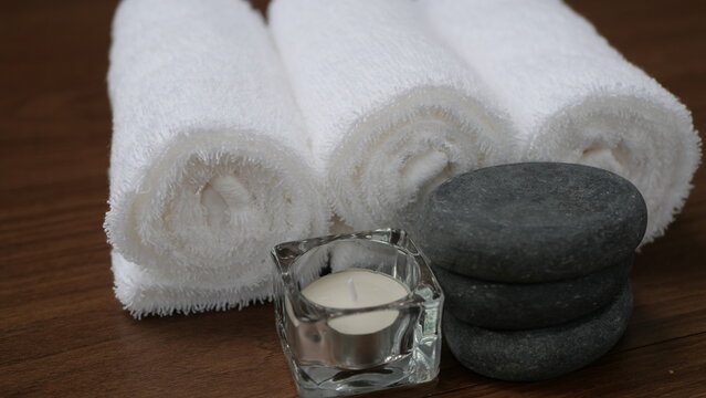 The image depicts a wellness spa atmosphere, decorated with white towels, black hot stones, and candles against a wooden background, conveying feelings of relaxation, massage therapy, tranquility, bea
