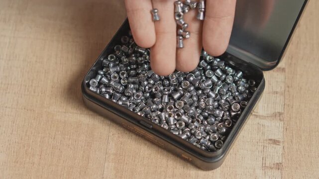 Hand taking and sorting shiny lead airgun pellets from open metal box on wooden table