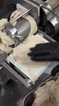 Close vertical view of black gloved hands guiding fresh corn masa through a stainless steel tortilla press that flattens dough into round tortillas inside a commercial kitchen