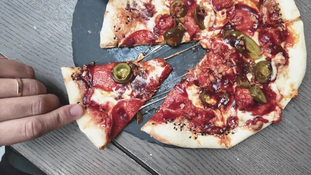 A person grabs a hot, cheesy slice of pepperoni jalape&ntilde;o pizza from a rustic wooden table in a close-up shot. The melted cheese stretches as the pizza slice is lifted, showcasing delicious detail.
