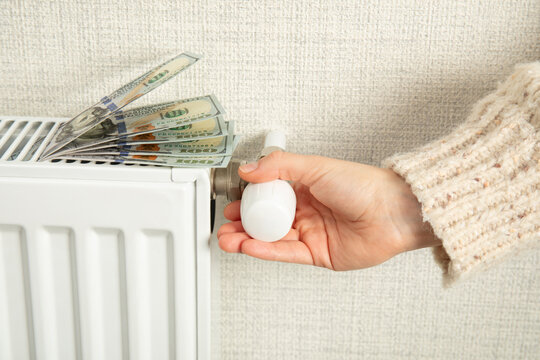 Heating season. Hand adjusts thermostat while money rests on radiator. Cost management concept
