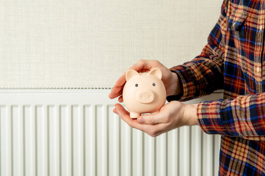 Heating season. Person holds piggy bank near radiator indicating savings during heating season