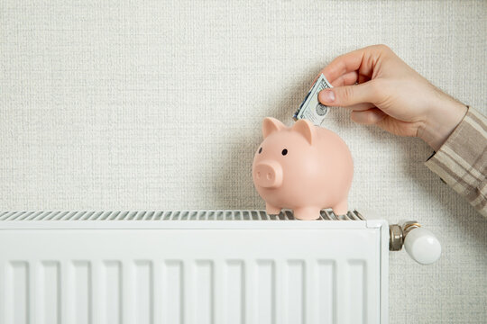 Heating season. Hand inserts cash into piggy bank placed on radiator
