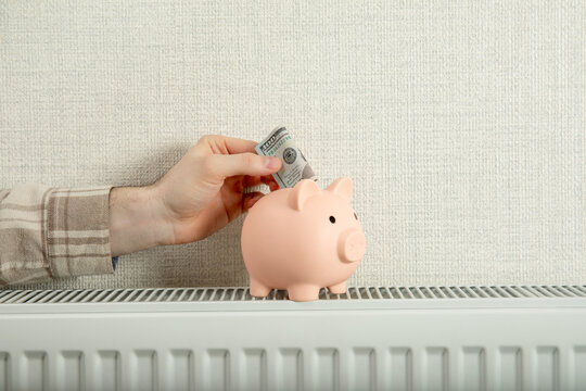 Heating season. Hand places money into piggy bank sitting on home radiator