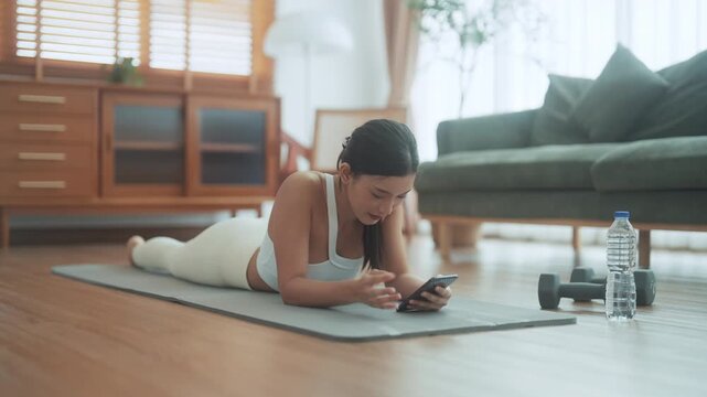 Asian woman resting after home workout and using mobile phone. Young sporty female after practicing yoga, break in doing exercise, relaxing on yoga mat, texting on the cell phone, holding smartphone