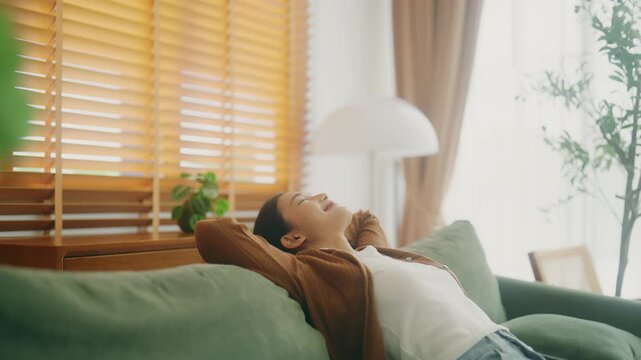 Relaxed young Asian woman enjoying rest on comfortable sofa, calm attractive girl relaxing on couch, breathing fresh air with eyes closed, meditating at home