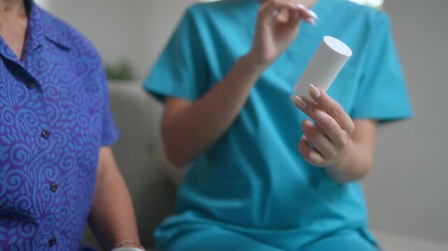 Close up of a female caregiver in scrubs holding a medicine bottle and explaining the prescription details and dosage to an elderly woman during a home visit or at a nursing facility
