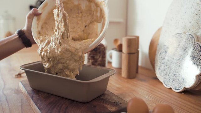 Batter is poured from a large mixing bowl into a rectangular loaf pan by a person wearing a dark bracelet who is working in a home kitchen on a wooden countertop near organic brown chicken eggs today.
