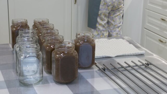 Filled glass jars sit neatly on a checkered table as a woman stands nearby in a bright kitchen. The scene highlights home preservation and a rewarding task. It feels peaceful and cozy.