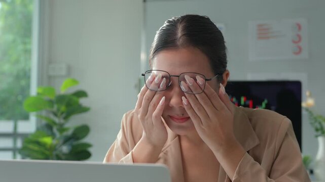 Stressed young asian businesswoman working late on a laptop, feeling exhausted and suffering from eye strain, rubbing her eyes and trying to concentrate on her work in the office