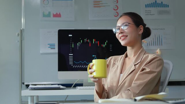 Young Asian businesswoman and financial analyst smiling and holding a yellow mug while sitting at her desk, with a candlestick chart displaying market data on the computer screen in the background
