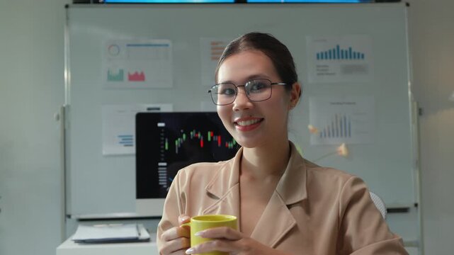 Confident young asian businesswoman holding a yellow mug, enjoying a coffee break in her modern office while looking at the camera with a cheerful smile and financial charts in the background