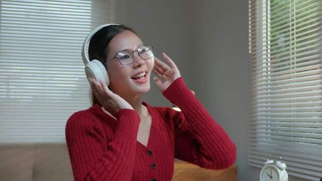 Happy young asian woman wearing headphones, listening to her favorite music, singing and dancing with pleasure in her cozy living room, enjoying a relaxing weekend at home