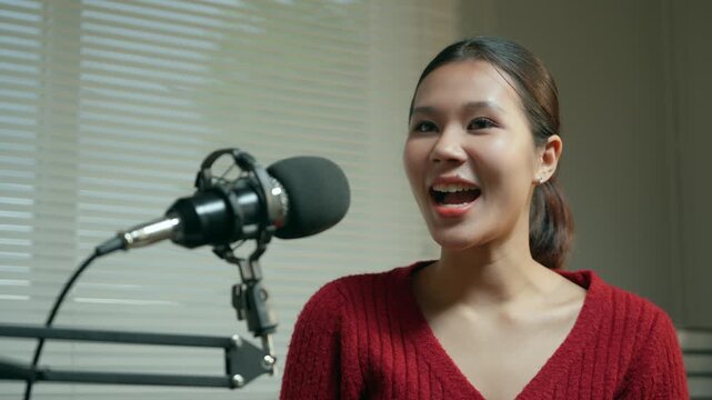 Confident Asian female podcaster speaking into a professional microphone during a live stream broadcast, sharing engaging content with her online audience from her home studio setup