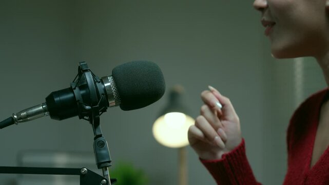Female radio host speaking into a condenser microphone and gesturing with her hands while recording a podcast or broadcasting live from her home studio in the evening
