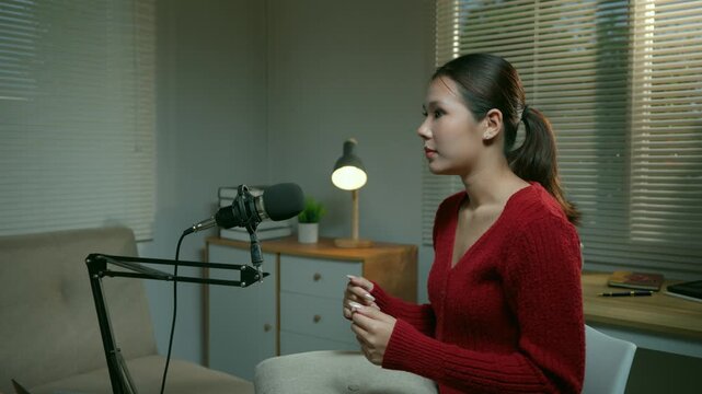 Young Asian woman enthusiastically speaking into a professional microphone, recording a live podcast or streaming content from her cozy home studio during the evening, engaging with her audience