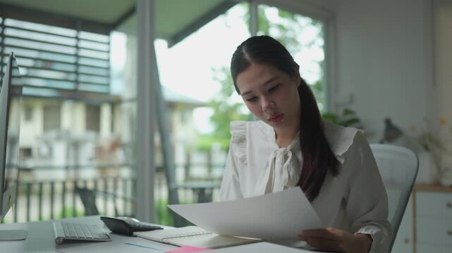 Concentrated asian businesswoman sitting at her desk, analyzing financial documents, calculating on a calculator, and taking notes while working on her computer in a modern home office