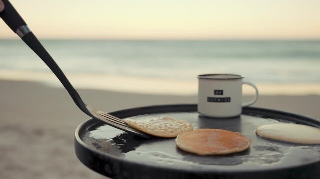 Pancake cooks on a black circular griddle placed on a sandy beach shore during sunset, with a metal camping mug sitting nearby while the gentle waves roll onto the beach in the blurred background.