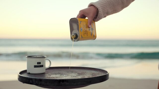 Woman pours oil from a small metal container onto a round portable camping stove while standing on the sandy beach at sunset, with a white enameled mug resting on the table near the calm ocean surf.