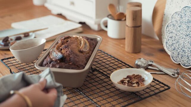 Banana bread loaf rests on a wire cooling rack after being pulled from the oven by a person holding a grey kitchen towel in a warm home kitchen environment with raw ingredients nearby on the table.