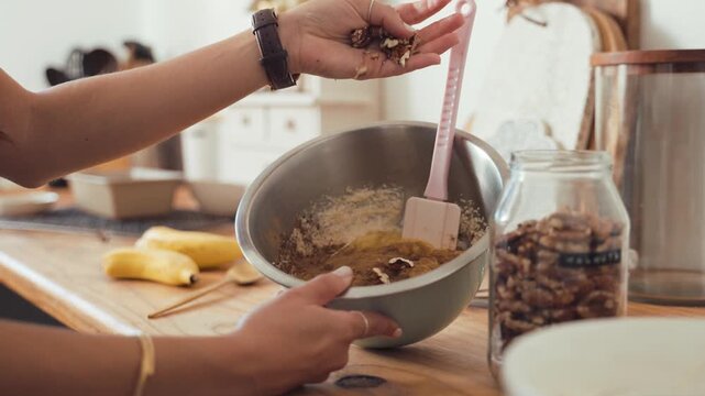 Woman adds crunchy pecans from her palm into a stainless steel bowl of cake batter while baking in a bright kitchen filled with various ingredients and jars on a warm wooden tabletop during the.