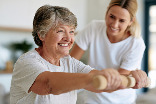 Warm light highlights two women. Senior woman smiles while exercising with a handle. Young woman supports her with a caring smile. Both wear casual white tops