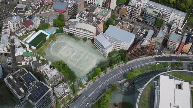 Aerial view of Tokyo, Japan, with dense urban buildings, curved roads, green trees and a large sports field. Modern cityscape for travel, business and urban planning themes.