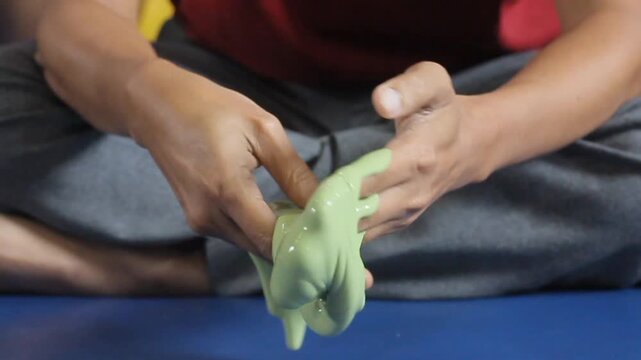 Hands stretch green slime with slow, focused motion. Fingers squeeze, pull, and twist the gooey material. Blue mat contrasts with the vibrant, textured substance. Sitting person appears calm