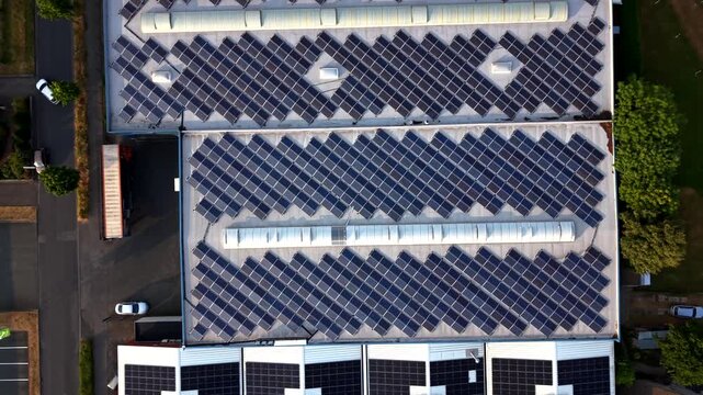 Solar panels blanket warehouse rooftops in downtown American district, parked cars and delivery trucks line adjacent street beside patches of green grass and shade trees. Aerial drone top down rise