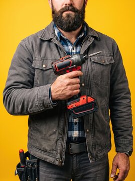 Bearded man in holds a cordless power drill, standing against a bright yellow background. Male hand and construction tools for house room renovation. Home renovation concept