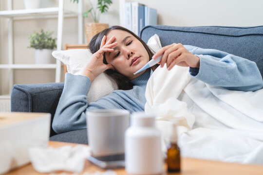 Sick person resting in bed checking temperature with thermometer, suffering from high fever, seasonal flu, influenza virus infection, and common cold symptoms at home