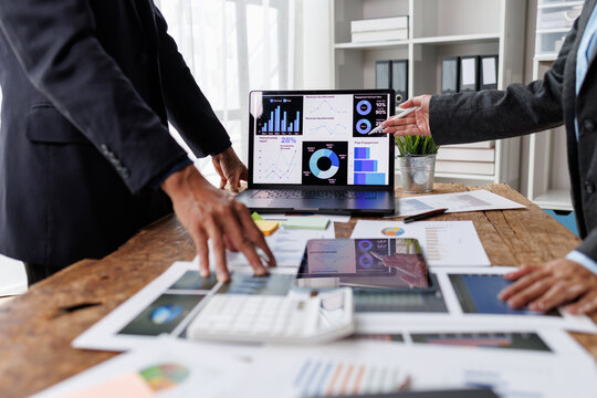Business professionals analyzing financial data dashboard on laptop screen and printed reports, collaborating on market strategy and performance statistics in an office meeting
