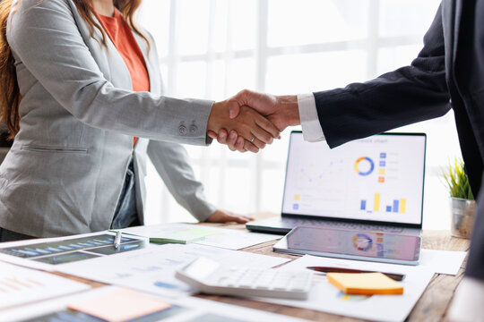 Two business professionals shaking hands over a desk filled with business documents and a laptop showing charts, illustrating a successful partnership and agreement in a corporate meeting