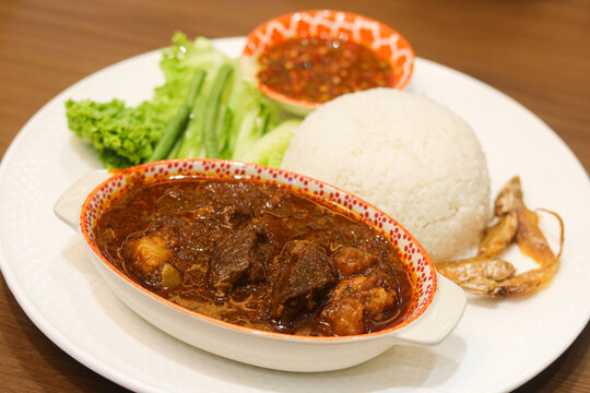 Sour and spicy beef stew served alongside a mound of steamed white rice, sambal chili, fresh vegetables, and salted fried fish.