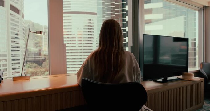 Woman typing on laptop at a desk in a modern high-rise office or hotel room overlooking a city skyline with skyscrapers during the day. Rear view medium shot. Slow motion.