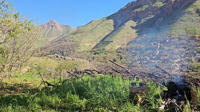 Small tea cups rest on a flat stone slab beside a blazing campfire with kettles, overlooking a terraced village and rugged mountain peaks under a clear blue sky.
📍 Kamalla village, Hawraman Takht
