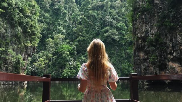 Young woman in floral dress standing on wooden balcony looking at tropical lagoon surrounded by limestone cliffs and lush jungle. Scenic travel destination medium shot from behind.