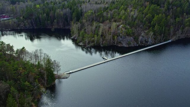 Aerial drone view of Stora Delsjon floating bridge crossing calm lake surrounded by dense forest in Delsjo nature reserve near Gothenburg Sweden. Water reflecting woodland creating peaceful scenery.