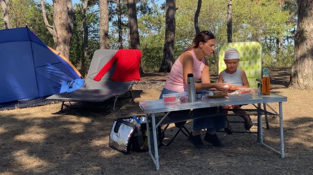 A loving mother and her small son eat a snack at a picnic table. They share a joyful meal under shady pine trees near a blue tent. This cozy camping day brings warmth and peace.