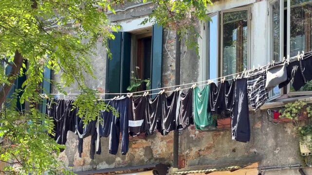 Static 4K shot of laundry drying on a clothesline outside a weathered house. Clothes sway gently in the breeze beneath leafy trees in bright daylight.