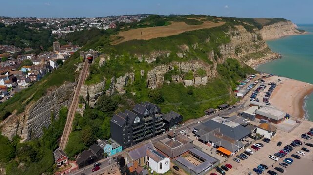 Hastings cliff funicular aerial push-in from Stade Beach toward East Hill, revealing chalky coastal cliffs, old town rooftops, promenade, and shingle shoreline on the English Channel.