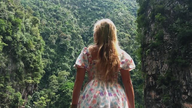 Blonde woman in floral dress looking at lush green tropical jungle valley between cliffs during daytime. Back view of traveler enjoying scenic landscape view in Southeast Asia. Slow motion.