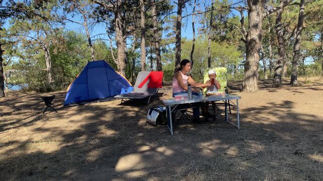 A loving mother sits with her happy son at a camping table as he drinks. They enjoy a sweet moment together. Sunny pine trees surround their peaceful campsite.