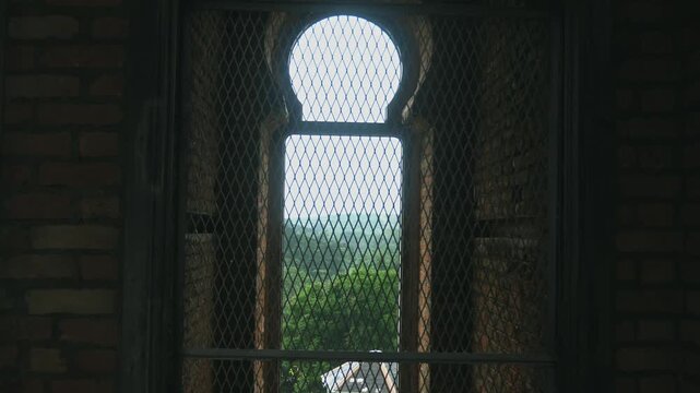 Arched stone window with metal grate overlooking green forest from dark ancient castle interior, historical architecture background, medieval heritage site, travel documentary