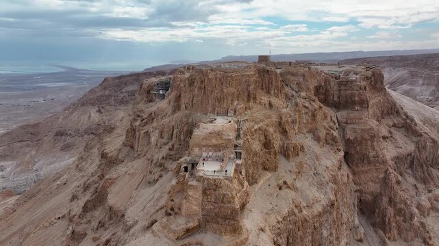 Drone tracking backward shot revealing the multi-tiered stone structures of the ancient mountaintop fortress with a wide panoramic overview of the Judean desert plain and salt sea under dramatic overc