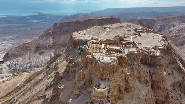 Drone tracking backward shot revealing the ancient stone fortress walls of Masada standing over a massive desert canyon valley under a blue sky during a sunny day.