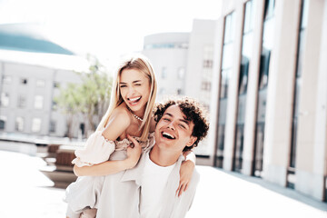 Obraz premium Young smiling beautiful woman and her handsome boyfriend in casual summer clothes. Happy cheerful family. Couple posing in street at sunny day. Female sits at his back. piggyback riding